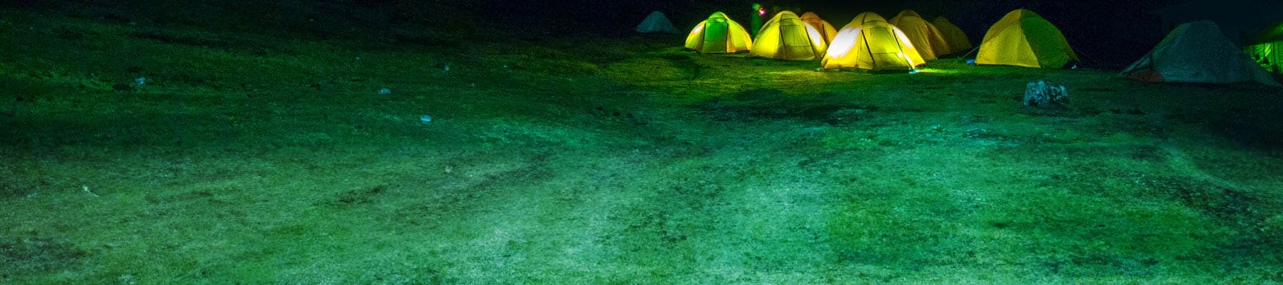 A starry sky framed by mountains. At the base of the mountains, several tents glow from lanterns lit within.